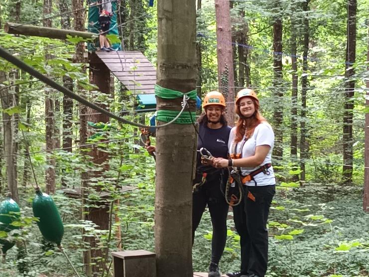 Zwei Personen in Sicherheitsgeschirr und -helmen stehen auf einer Plattform im Wald, während ein dritter Teilnehmer sich vorbereitet.