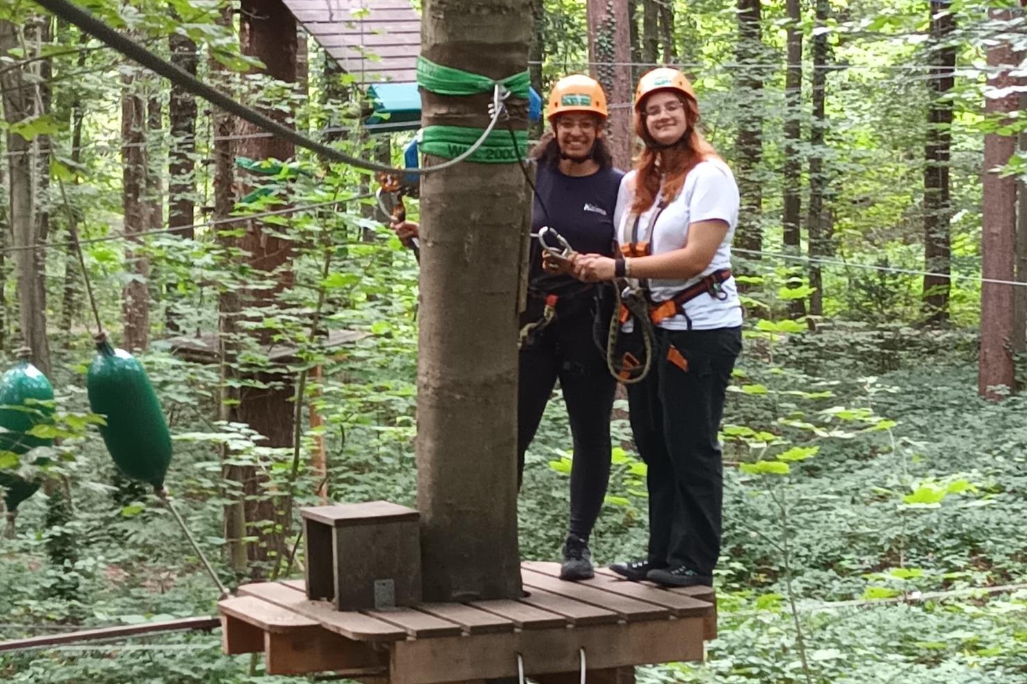Zwei Personen in Sicherheitsgeschirr und -helmen stehen auf einer Plattform im Wald, w&auml;hrend ein dritter Teilnehmer sich vorbereitet.