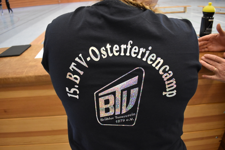 R&uuml;ckansicht eines schwarzen T-Shirts mit dem Aufdruck &bdquo;15. BTV-Osterferiencamp&ldquo; und dem Logo des Br&uuml;hler Turnvereins.