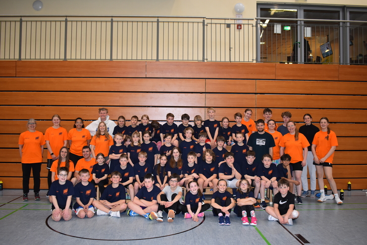 Gruppenshot von Kindern und Trainern in Sportkleidung in einer Halle, mit Holzw&auml;nden im Hintergrund.