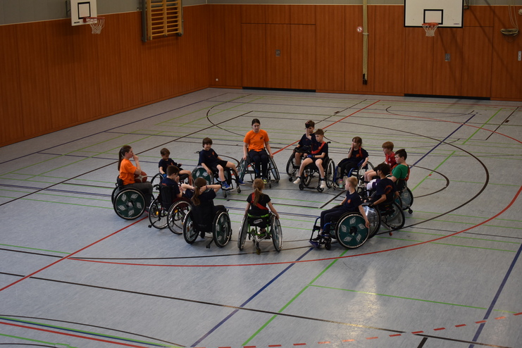 Gruppe von Kindern im Rollstuhl sitzt im Kreis in einer Sporthalle, umgeben von Linien auf dem Boden.