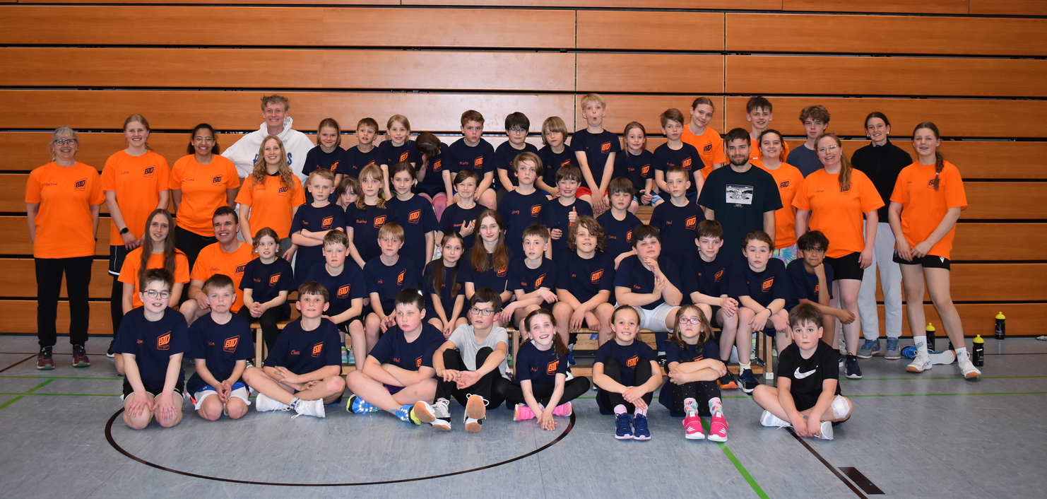 Gruppenshot von Kindern und Trainern in Sportkleidung in einer Halle, mit Holzwänden im Hintergrund.