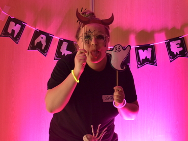 Frau mit Teufelshörnern und Gesichtsbemalung hält eine Geisterfigur, mit Halloween-Dekoration im Hintergrund.