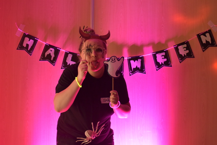 Frau mit Teufelshörnern und Gesichtsbemalung hält eine Geisterfigur, mit Halloween-Dekoration im Hintergrund.