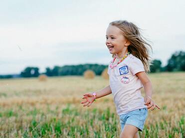 Lachendes kleines Mädchen mit langen Haaren, das durch ein Feld mit Heuballen läuft.