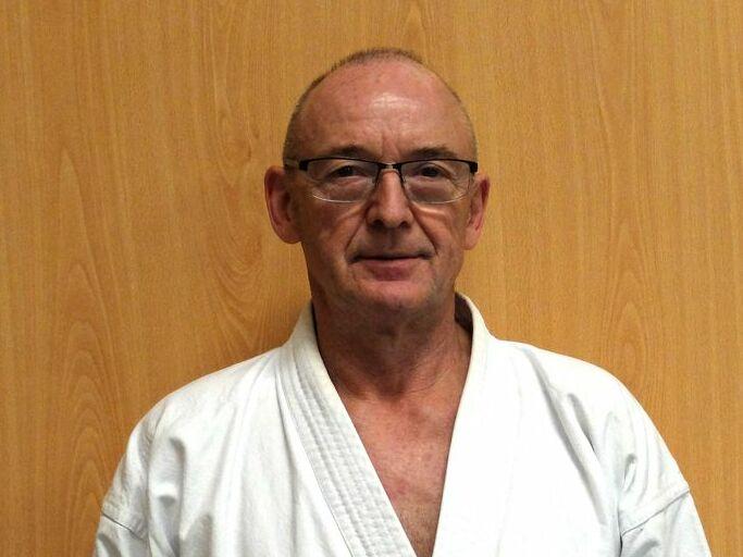 Ein älterer Mann in einem weißen Karateanzug mit schwarzem Gürtel steht vor einer Holzpaneel-Wand.