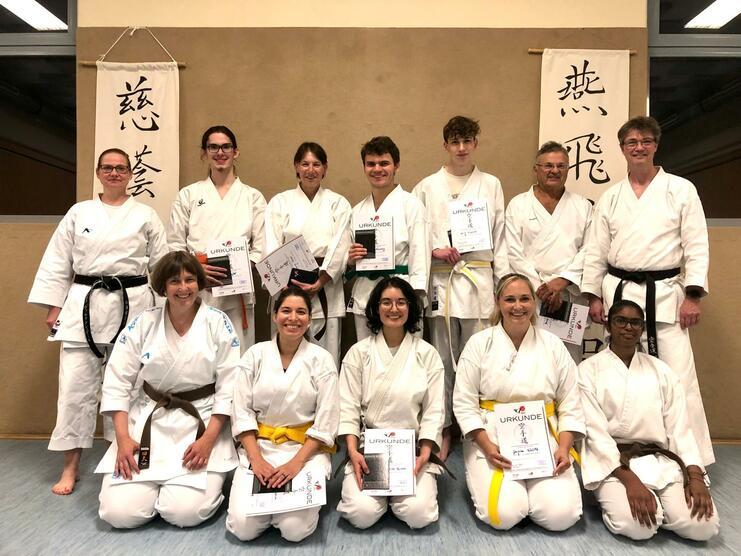 Gruppe von Karateka in Weiß mit Urkunden, posierend vor Wand mit Schriftzügen, inklusive Schwarz- und Braungurten.