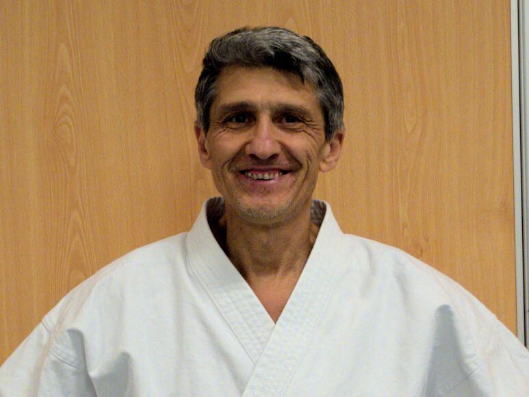 Mann in Karate-Uniform mit schwarzem Gürtel steht lächelnd vor einer Holzwand.