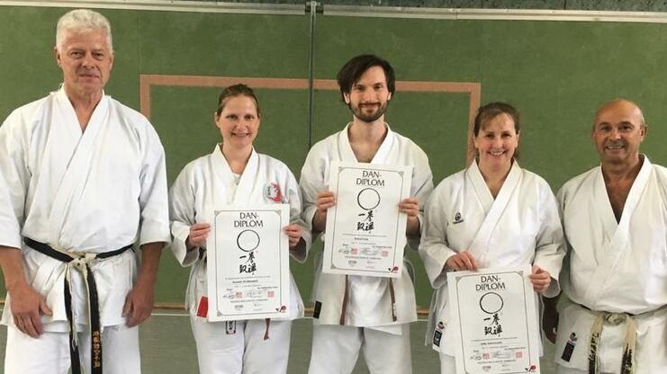 Gruppierung von vier Karateka in weißen Gi, die stolz ihre Dan-Urkunden in einer Sporthalle zeigen.