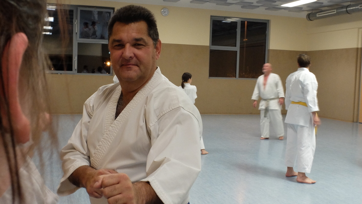 Ein Karatekämpfer in Weiß mit blauer Gürtelschärfe posiert in einem Trainingsraum, während andere sich im Hintergrund bewegen.