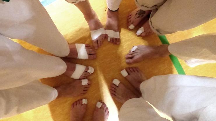 Sicht auf mehrere Füße in weißen Anzügen, eingewickelt in Bandagen, auf einem Hallenboden mit linierter Oberfläche.