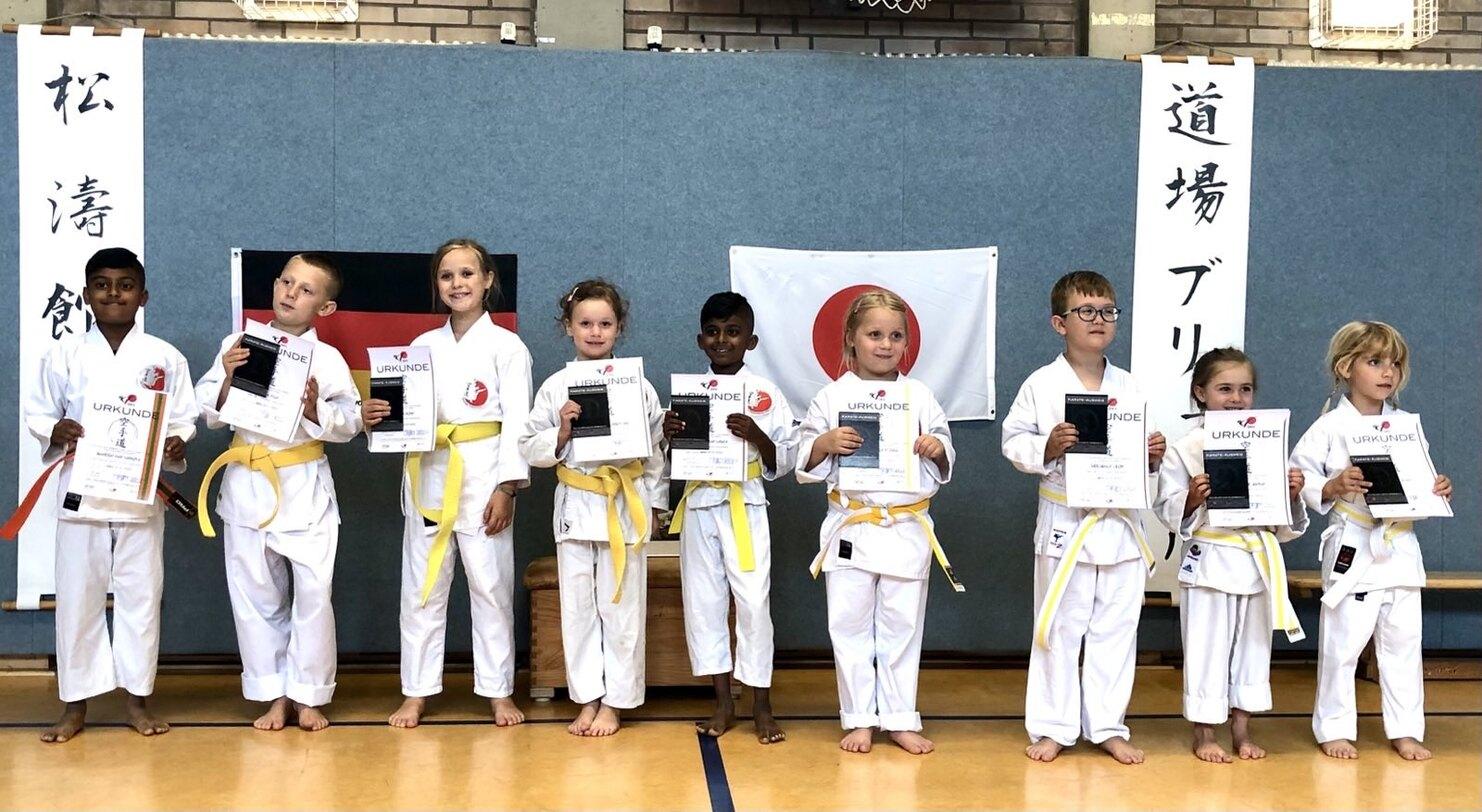 Gruppe von acht Kindern in Karateanzügen mit Urkunden in der Hand, vor einer blauen Wand mit Flaggen und Schriftzeichen.