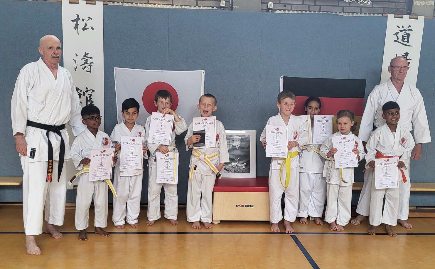 Gruppierung von Kindern in Karate-Uniformen mit Urkunden, zwei Trainern im Hintergrund, vor japanischer und deutscher Flagge.