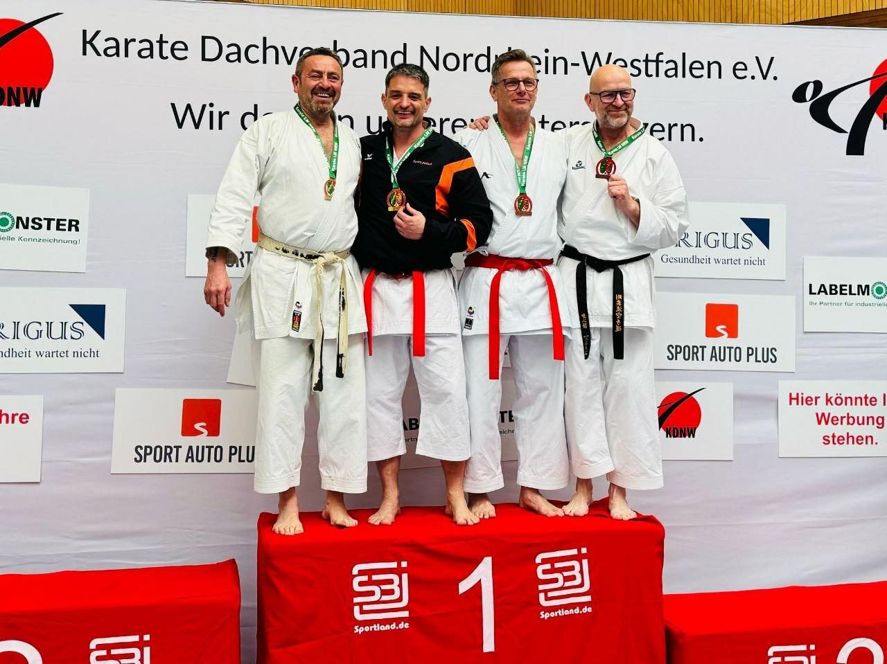 Vier Karateka in Weiß und Schwarz stehen auf einem Siegerpodest mit Medaillen, im Hintergrund das Logo des Karate Dachverbands NRW.