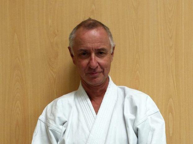 Mann in einem weißen Karate-Gi steht vor einer hellen Holzoptik-Wand, mit gefalteten Händen.
