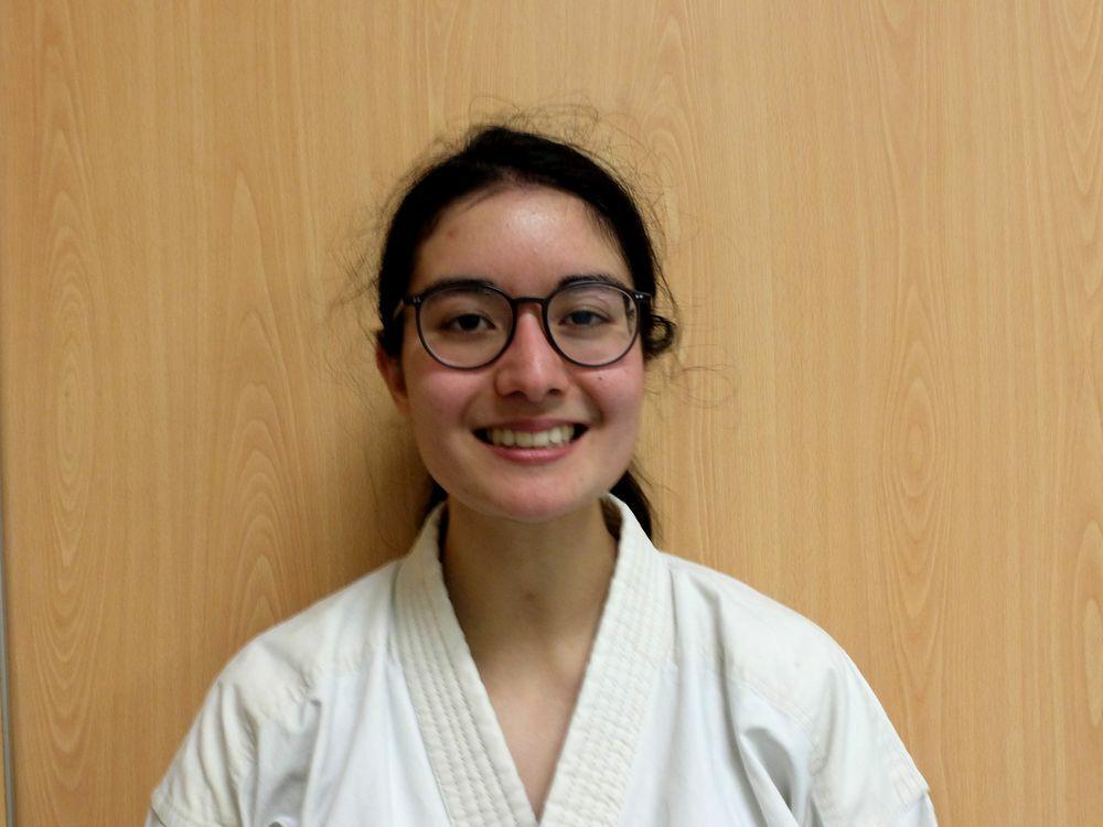 Junge Frau mit Brille und Karateanzug lächelt vor einer holzfarbenen Wand.