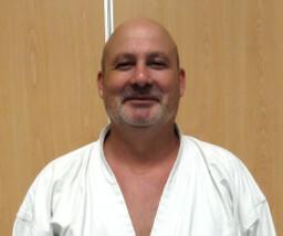 Mann in Karate-Uniform lächelt vor einer Holzpaneel-Wand.