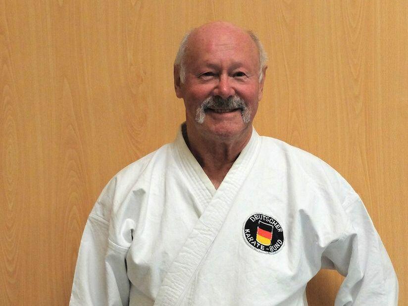 Älterer Mann in einem weißen Karate-Gi mit schwarzem Gürtel, lächelt vor einer holzfarbenen Wand.