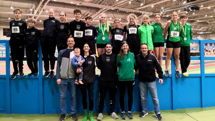 Gruppe von Sportlern in Sportkleidung, einige mit Medaillen, posieren lächelnd auf einer Sportanlage.