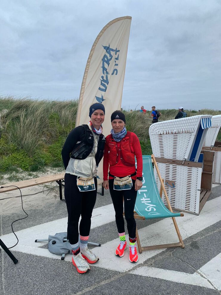 Zwei L&auml;uferinnen posieren vor einem "Ultra"-Banner am Strand, mit Laufbekleidung und bunten Socken.