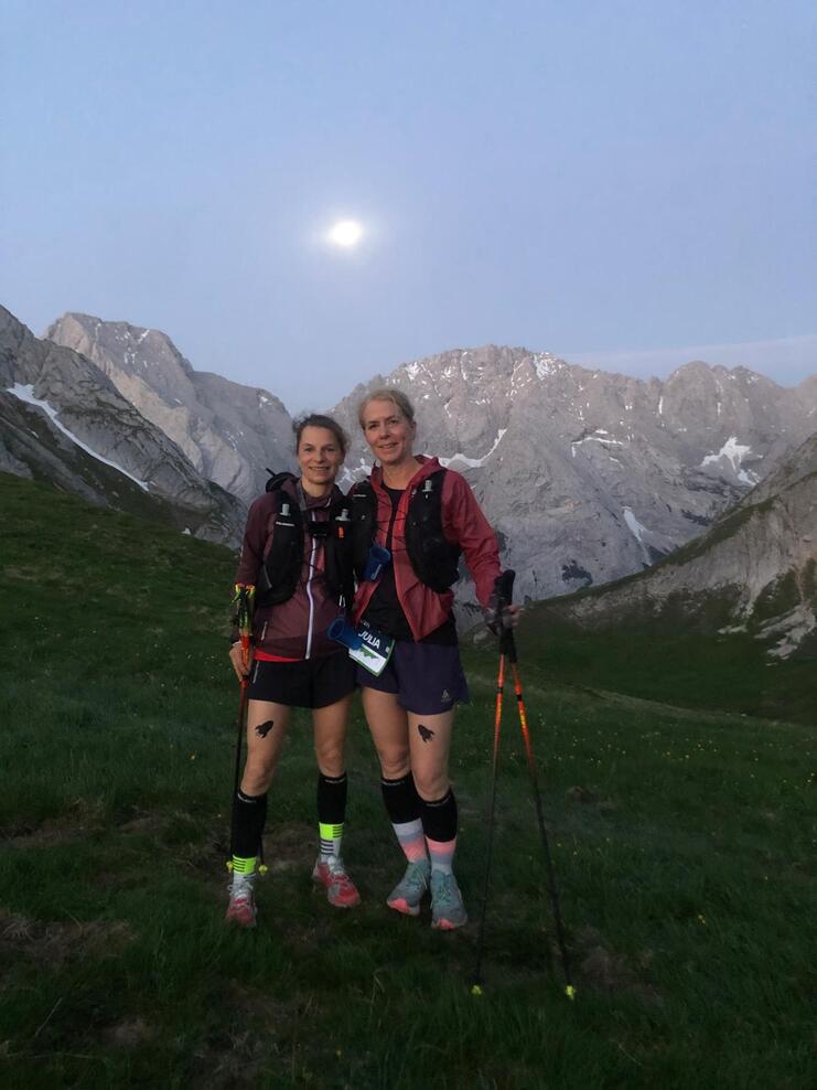 Zwei Frauen stehen auf einer Wiese in den Bergen, beide mit Wanderstöcken und Sportkleidung, der Mond ist sichtbar.