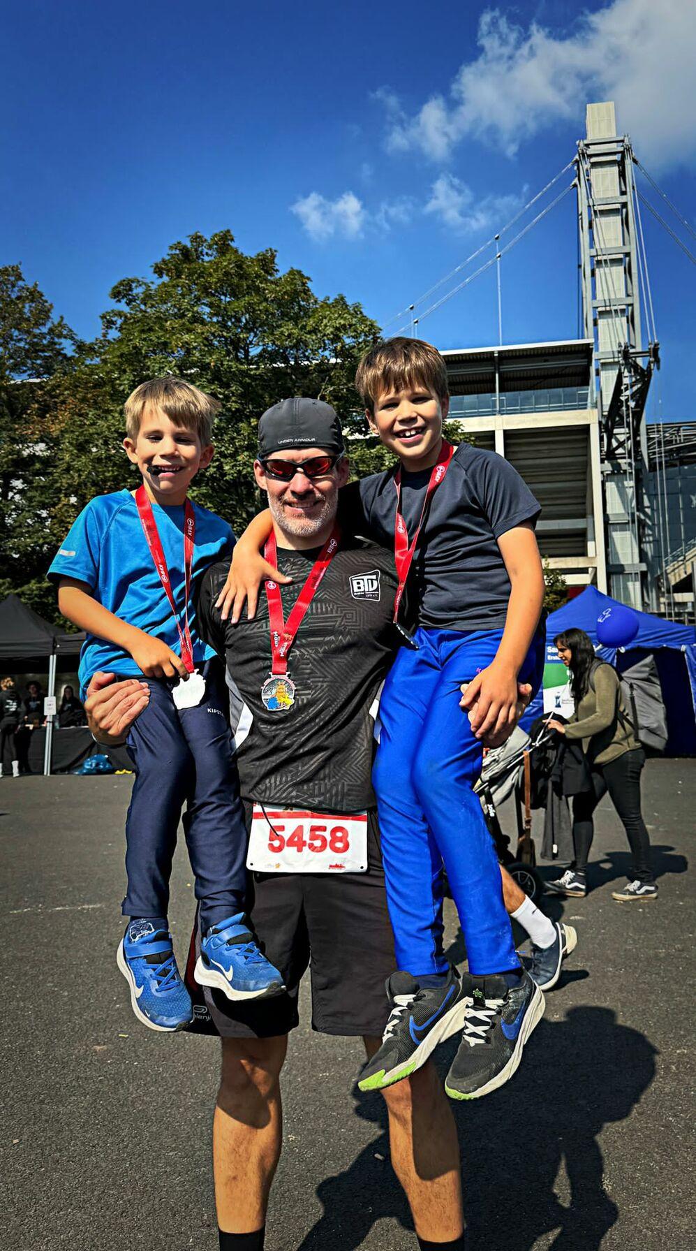 Ein Mann steht auf einem Parkplatz und trägt jeweils einen Jungen auf seinen Armen, beide mit Medaillen um den Hals.
