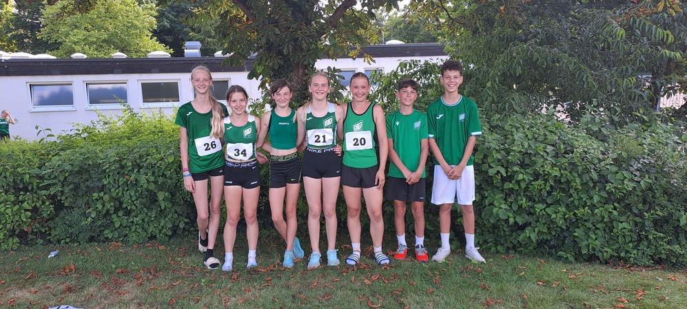 Gruppe von sieben Jugendlichen in sportlicher Kleidung, posierend vor einer Hecke und einem Gebäude.
