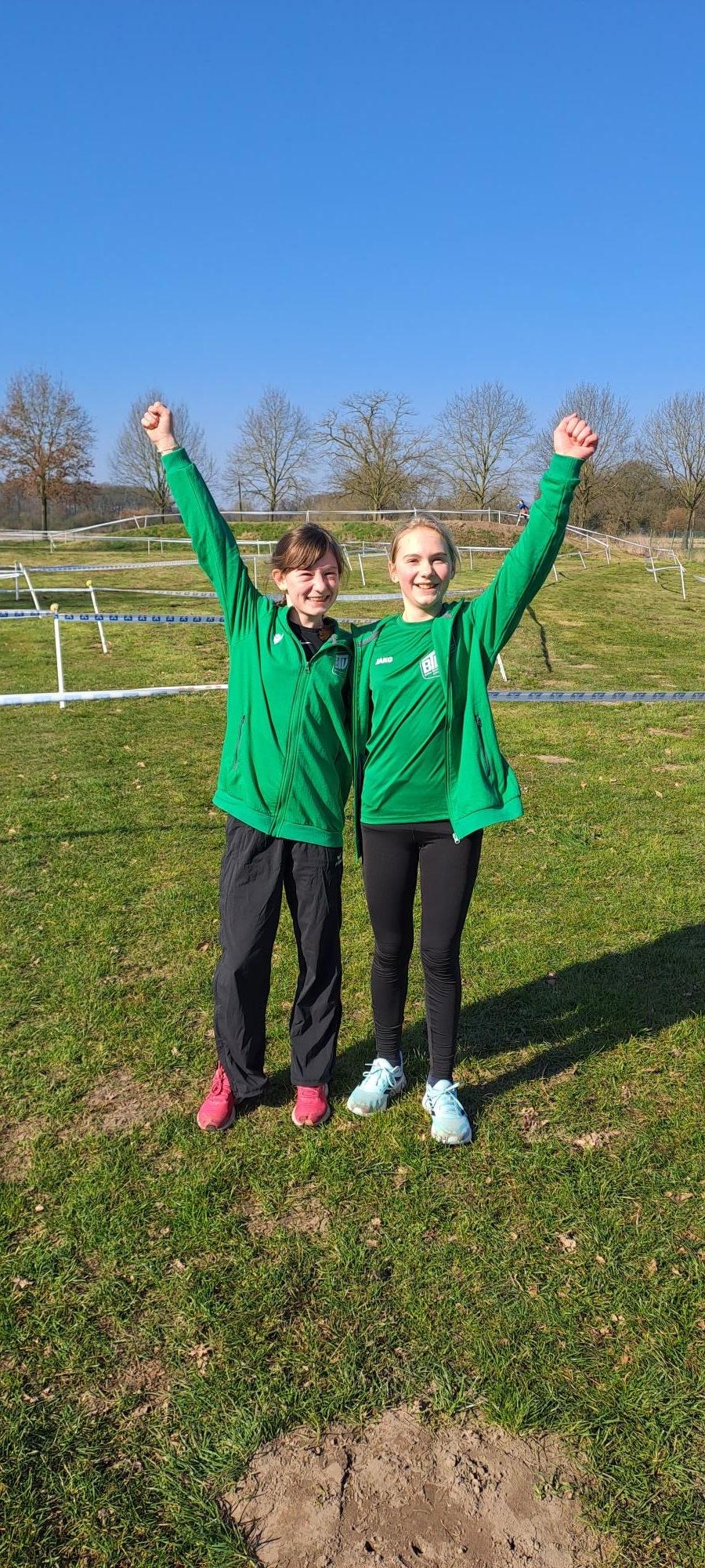 Zwei Mädchen in grünen Sportjacken stehen im Freien und heben die Arme in Freude. Hintergrund mit vereinzelten Bäumen.
