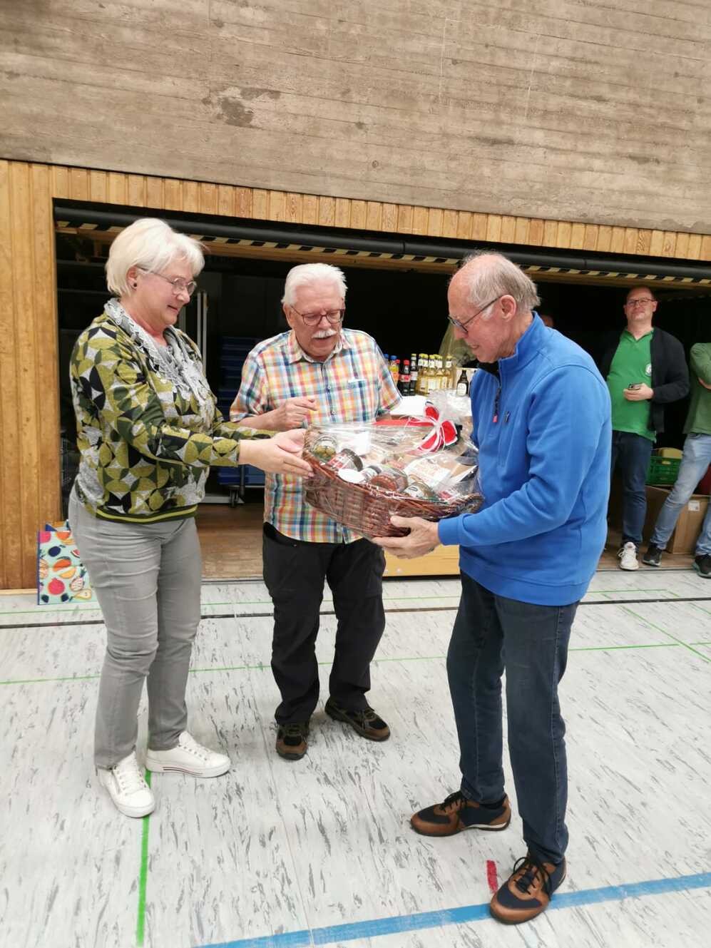 Drei ältere Personen überreichen sich einen Korb mit Geschenken in einer Sporthalle. Im Hintergrund sind Getränke sichtbar.