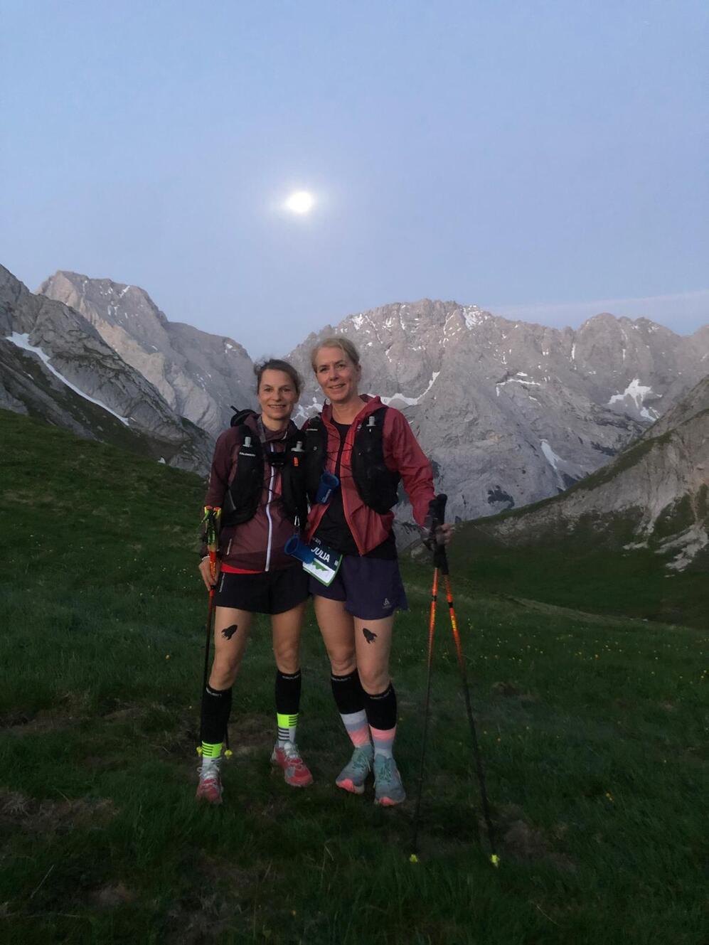 Zwei Frauen stehen auf einer Wiese in den Bergen, beide mit Wanderstöcken und Sportkleidung, der Mond ist sichtbar.