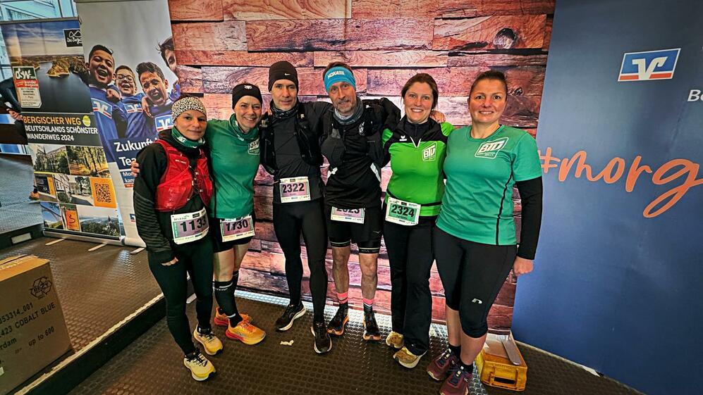 Gruppe von sechs Läufern in Sportkleidung mit Startnummern vor einer Holzpaneel-Wand und einem Werbebanner.