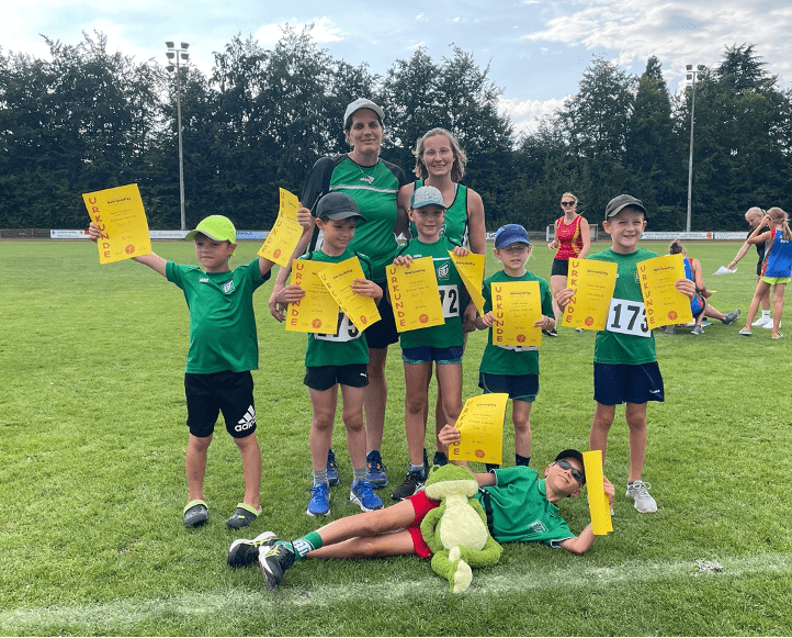 Gruppe junger Athleten in grünen Outfits hält Urkunden auf einem Sportplatz, ein Kind liegt mit Teddybär auf dem Rasen.