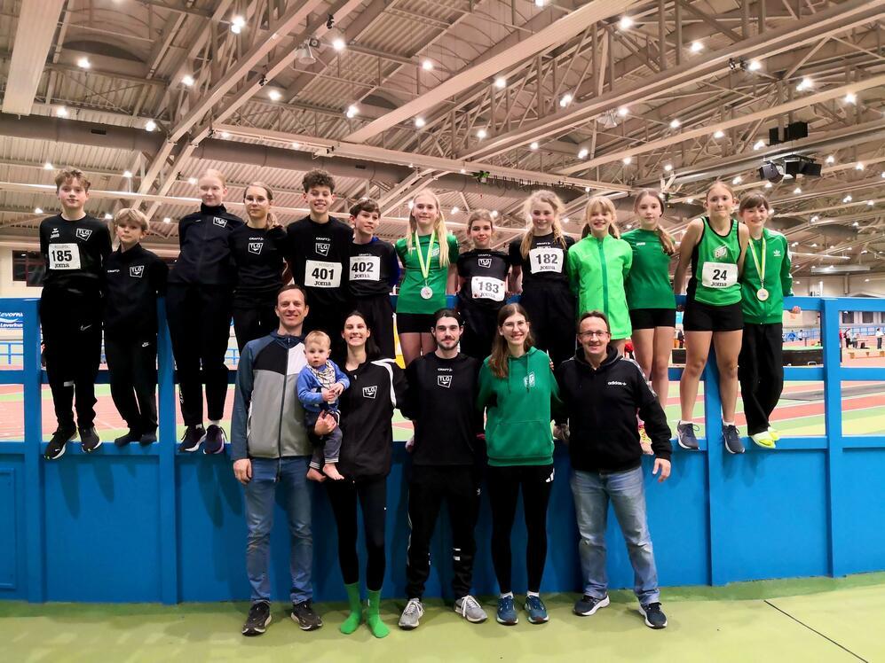 Gruppe von Sportlern in Sportkleidung, einige mit Medaillen, posieren lächelnd auf einer Sportanlage.