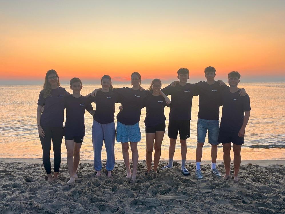 Gruppe von acht Jugendlichen in T-Shirts am Strand vor einem Sonnenuntergang, Barfuß im Sand stehend.