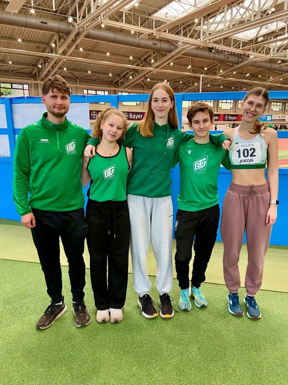 Fünf junge Athleten in grünen Trainingsoutfits stehen lächelnd auf einem Sportplatz unter einer hohen Decke.