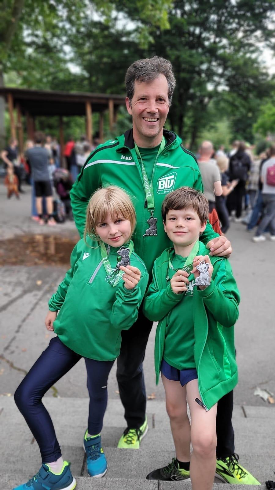 Ein Mann mit zwei Kindern steht vor einer Menschenmenge und hält Medaillen in den Händen, alle tragen grüne Kleidung.