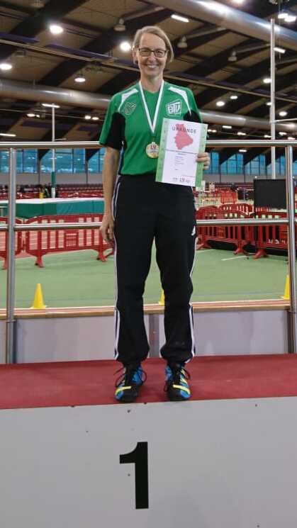 Sportlerin auf dem Siegertreppchen, Medaille und Urkunde in der Hand, in einer Sporthalle mit grünem Boden.