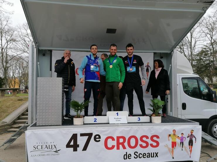 Drei Medaillengewinner stehen auf dem Podium eines Wettbewerbs, während ein Mann eine Ansprache hält.
