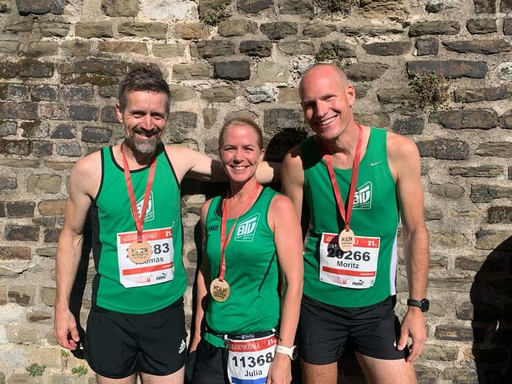 Drei L&auml;ufer in gr&uuml;nen Trikots mit Medaillen stehen vor einer alten Steinmauer, l&auml;chelnd nach einem Wettkampf.