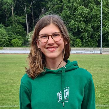 Junge Frau mit lächelndem Gesicht, trägt grünes Hoodie, hinter ihr ist ein Sportplatz mit Bäumen im Hintergrund.