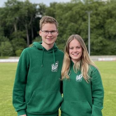 Zwei Jugendliche in grünen Hoodies stehen freundlich nebeneinander auf einem Sportplatz.