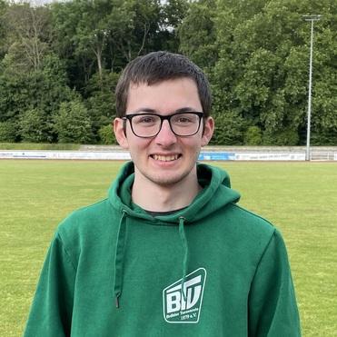 Junger Mann in grünem Hoodie mit Vereinslogo lächelt vor einem Sportplatz mit Bäumen im Hintergrund.