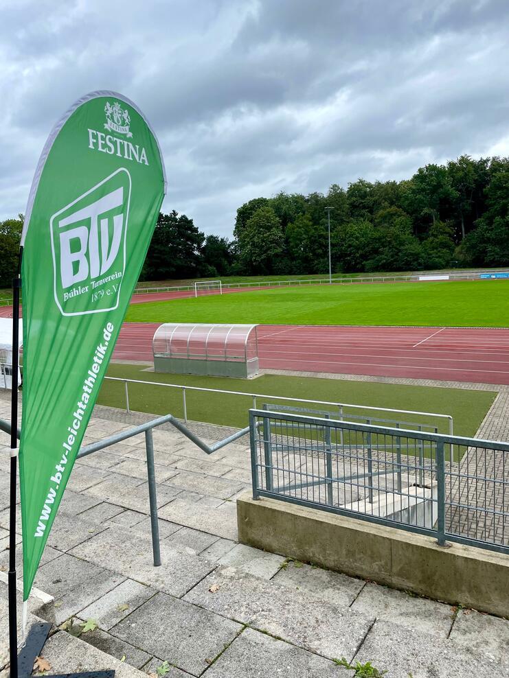 Grünes Werbebanner des Bühler Turnvereins an einer Sportanlage mit Fußballfeld und Rasentribüne im Hintergrund.