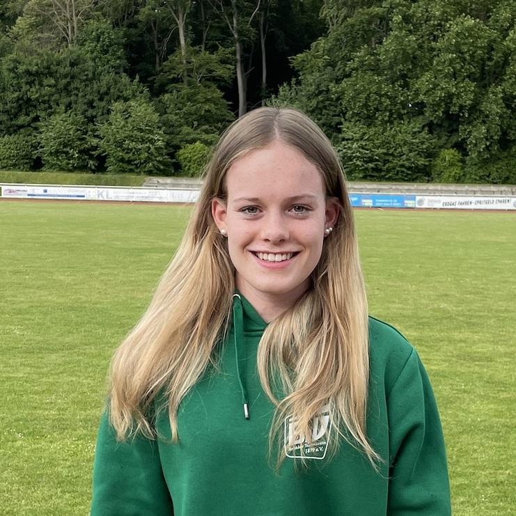 Junge Frau mit langen blonden Haaren und grünem Hoodie lächelt auf einem Sportplatz mit Rasen im Hintergrund.