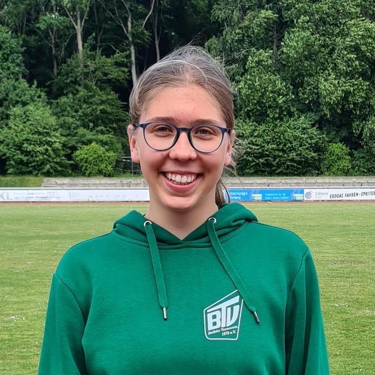 Junge Frau mit Brille und grünem Kapuzenpullover lächelt vor einer Sportplatzkulisse mit Bäumen im Hintergrund.