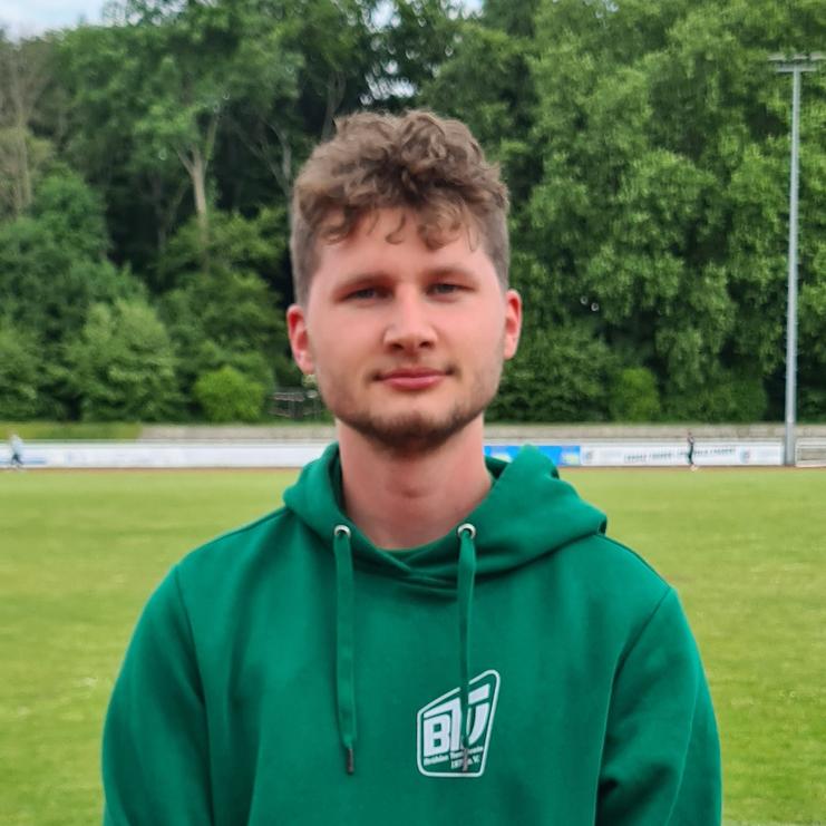 Junger Mann in grünem Kapuzenpullover steht auf einem Sportplatz, umgeben von Grünanlagen.