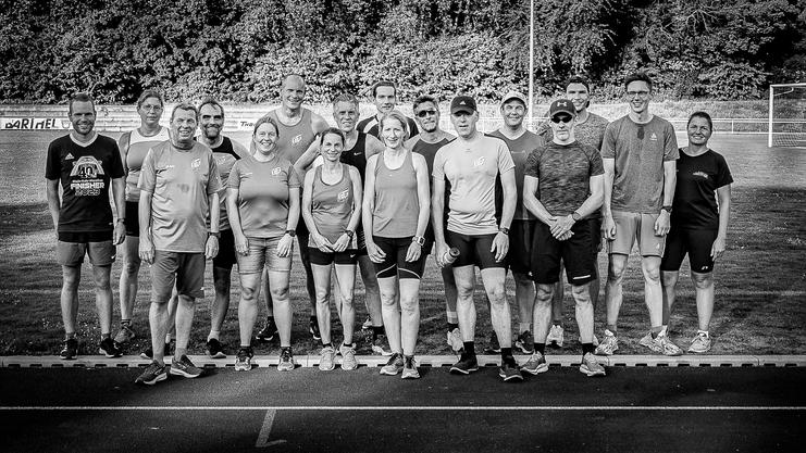 Gruppe von 16 Sportlern in Sportkleidung steht auf einer Laufbahn, umgeben von grüner Umgebung. Foto in Schwarzweiß.