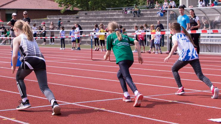 Vier Kinder starten beim Hürdenlauf auf einer roten Laufbahn, während Zuschauer im Hintergrund zuschauen.