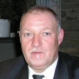 Ein Mann mit kurzen, grauen Haaren und einem Anzug posiert in einem Innenraum. Hintergrund schwach erkennbar.