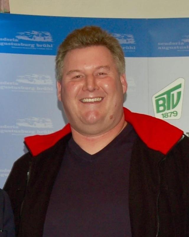 Ein lächelnder Mann mit kurzen, grauen Haaren in einem schwarzen und roten Oberteil vor einem blauen Hintergrund.